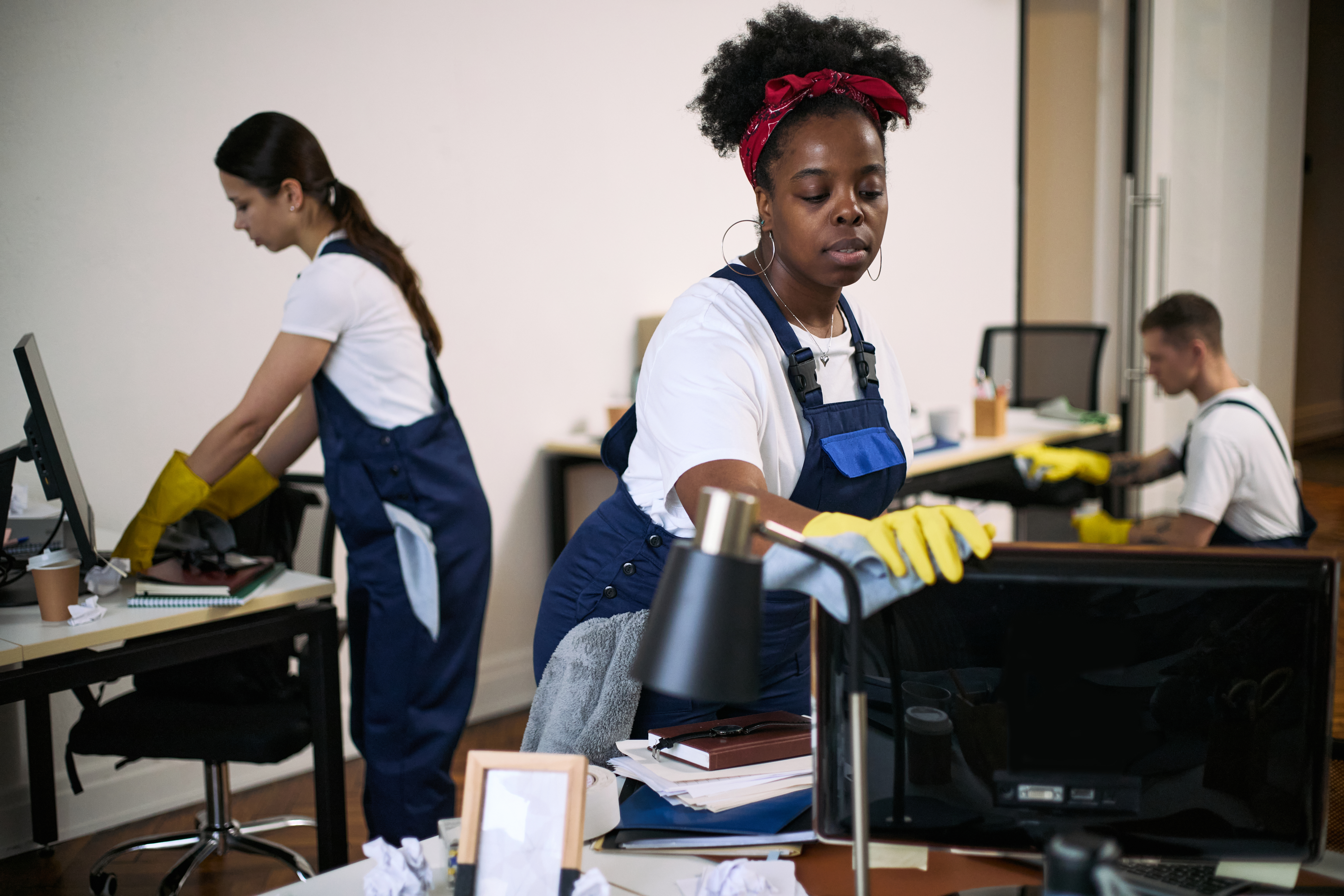 Black young adult woman cleaning computer monitor with cloth and gloves in modern office, while Caucasian young adult woman and Caucasian young adult man cleaning desks in background